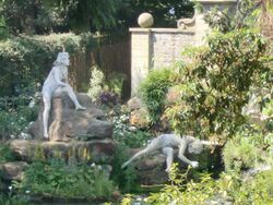 Colour photograph, zoom shot through trees of two nude female statues on a rockery, one sitting, the other in a strange pose