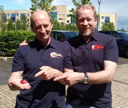 Tim (right) and Chris (left) Stamper; the middle-aged men both wear black polos with red logos on the right of the buttons. Tim has a beard, while Chris is balding.