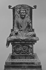 Frontal view of a cross-legged seated statue placed on a throne. His right arm rests on his leg, the left arm is bent and slightly raised.