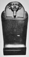 Black and white image of a dark stone coffin with a human face, the coffin stands upright facing the viewer.