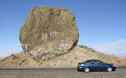 This photo shows an automobile passing in front of a rock which is essentially fully exposed. The rock has a rough, dark surface indicating it is weathered basalt and is roughly circular in exposed cross-section. The rock is immediately adjacent to a roadway—the road cut removed much of the earth from one side of it exposing it—from the excavation it is evident that the rock sits on a mound of glacial till. The rock is approximately two times the length of the car (i.e., ≈9 metres) in one direction and five times the height of the car in the other direction (i.e., ≈9 metres). Since the rock has not tipped onto the road and no structural support is provided, it must be approximately as deep as it is wide and high. Since the density of basalt is 3 grams per cubic centimetre, this puts the mass of the rock at about 400 to 500 metric tons (consistent with the references).
