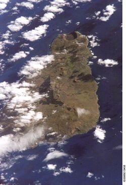 A colour photo from space showing a brownish triangular island in a dark blue sea; all partially obscured by white clouds especially on the left.