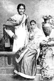 Black-and-white photograph of a finely dressed man and woman: the man, smiling, stands with the hand on the hip and elbow turned outward with a shawl draped over his shoulders and in Bengali formal wear. In front of him, the woman, seated, is in elaborate dress and shawl; she leans against a carved table supporting a vase and flowing leaves.
