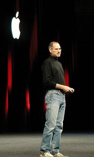 Full-length portrait of a middle-aged man, wearing jeans and a black turtleneck shirt, standing in front of a dark curtain with a white Apple logo
