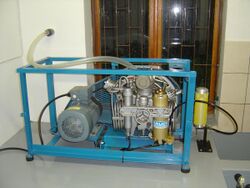 A small high-pressure compressor mounted on a steel frame with a three-phase electric motor for power. A flexible plastic air intake hose provides fresh air from outside of the building.