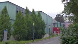 Street leading to the red entrance of the Max Planck Institute for Gravitational Physics in Hannover. The building is partly hidden behind trees next to the street.