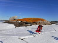 Red backpack resting on snow in front of a large orange-stained rock under a clear blue sky.