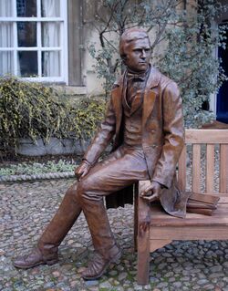 Bronze statue of Darwin in 1830 clothes, seated on the arm of a wooden bench; behind him plants partly cover a stone wall, a window has white-painted wooden frames