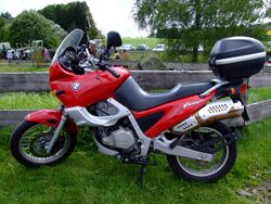 Red BMW F650ST with topbox parked on grass in front of a rustic wooden fence