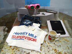 An image of the personal belongings of Harvey Milk. Visible in the photo are Milk's battered, gold-painted kitchen table; a "Harvey Milk/Supervisor" campaign t-shirt; a pair of his Levi's jeans; his appointment book for 1977; his personal-safety whistle; his harmonica; and a pair of pink novelty sunglasses owned by Milk. Also on display is a mug owned by Scott Smith.