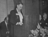 A candid photograph of a man wearing a suit speaking at a lectern