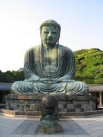 Front view of an outside cross-legged seated statue. The eyes are closed and the hands rest on the lap with palms facing upward. The statue is left-right symmetric, appears green of copper rust and is placed on a stone platform.