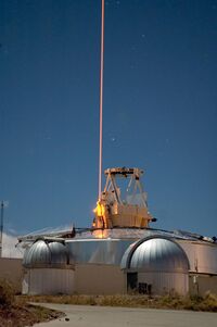 A thin bright yellow light beam goes straight up into the sky from an optical instrument