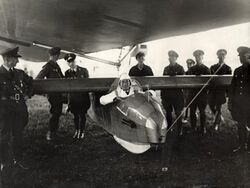 A black-and-white photograph of a man sitting in a small, polished-wood sports glider, underneath the bulk of the zeppelin. He is wearing a white flight suit, and a white fabric flight helmet. His elbows are resting on the sides of the cockpit. Behind and around him are nine men in military officers' dress uniform. The glider is secured to the airship by a cable at its nose and a mechanical attachment at its centre.