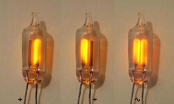 Photograph of 3 small glass capsules. Each capsule has 2 parallel wires that pass through the glass. Inside the left capsule, the right electrode is glowing orange. In the middle capsule, the left electrode is glowing. In the right capsule, both electrodes are glowing.