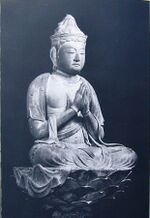 Three-quarter view of a cross-legged seated deity on a pedestal with hands joined as in prayer in front of the body.