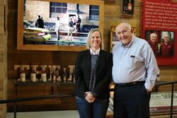 A caucasian known as Jenny Murphy in jeans, a navy long sleeve, a white collared undershirt, and a black blazer is standing next to Jimmy Russel, a taller caucasian man who is bald on top, wearing black pants and a light blue button-up shirt. Both individuals are standing before history memorabilia at the Wild Turkey Distillery, including several bottles and older photographs.