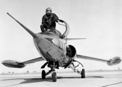 Pilot in overall black suit crouching on jet aircraft's nose