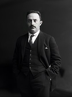 A black-and-white portrait of a formally&dressed young man with a short, black mustache wearing a light-colored hat, white shirt, a light-colored suit, a dark tie, and dark shoes. The man is outside a building from which a dog is coming out.