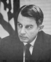 A photograph of a man wearing a suit looking towards the left with a flag behind him