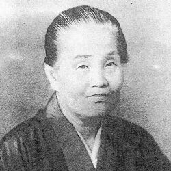 An older Japanese woman, hair dressed back from her face, wearing a dark kimono