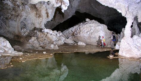 Two cavers inside of a cave look at the roof of the cave while standing at the shore of an underwater body of water.