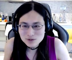A woman with long black hair, black headphones and dark wire-rimmed glasses looks at the camera. She is wearing a sleeveless purple top and choker-length necklace and is sitting in a gamer chair.