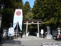 An image of the entrance of Kumano Hongu Taisha, a torii gate stands prominently in front of tall green trees. To the side are Japanese paper lanterns, and a prominent banner stands in front of a three-legged-crow.