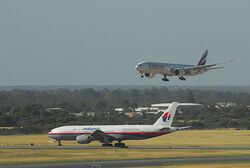 A white Emirates aircraft above runway, with landing gear down, and a Malaysia Airlines jet in red, white, and blue livery taxiing