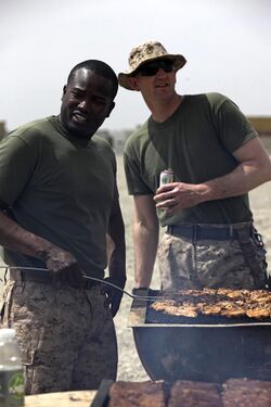 Two men in identical short-sleeved shirts and camouflage pants, one very dark-skinned with no hat and one very light-skinned wearing a hat and sunglasses, stand smiling over a barbecue full of cooking meat in a bright location.