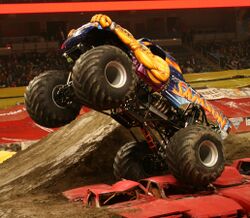 A suspended Samson crushes automobiles in the 2011 Monster Jam monster truck event. The floor of the venue is covered in dirt, and a dirt ramp is adjacent to the right side of Samson. Objects in background include crowd-filled stands and promotional images.