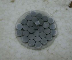 Dark grey pills on a watchglass. One cubic piece of the same material on top of the pills.