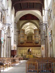 An interior view of the nave of Rochester Cathedral, looking towards the east. The nave has Norman arches and a flat wooden ceiling, beyond which there the stone vault of the choir. The cathedral is divided by a stone screen on which rest two section of richly decorated organ pipes.