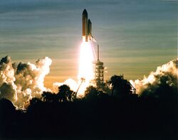 A space shuttle launches into a dawn sky. Clouds in the sky, in the launch plume and from the flame trench, are visible, as is the scaffolding-like launchpad and some vegetation silhouetted in the foreground.