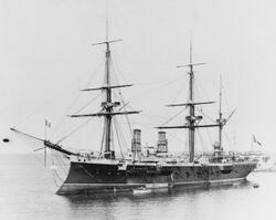 A large black ship with three masts and sail rigging sits in a harbor with several small boats alongside.