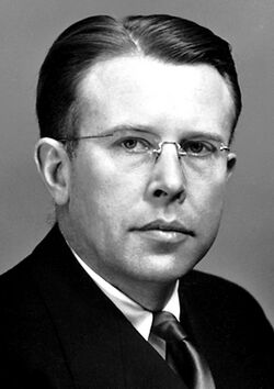 Head and shoulders of a man wearing rimless glasses, and a dark suit and tie