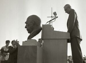 Oppenheimer stands behind a large bust and gives a speech