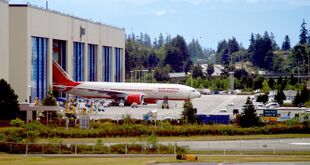 Aircraft factory. Rectangular building with six doors, one of which is open to reveal an emerging airliner. The background is a forest; small vehicles and taxiways surround the factory.