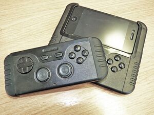 Two game control devices sit atop one another. The upper is a gamepad alone, while the lower is attached to a phone.