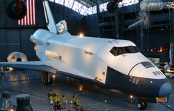 Enterprise on display in a black hangar filled with other space artifacts including the tops of Mercury and Gemini capsules, rockets and satellites, a Manned Maneuvering Unit, and other space artifacts. An American flag hangs on the wall of the hangar in the back. The Shuttle is resting on its landing gear with the payload bay doors closed, and museum visitors are kept away by stanchions. 13 visitors are visible in this picture, one group is wearing matching yellow shirts. There is ample room for more visitors.