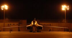 Front view of B-1 parked on ramp at night. Nearby yellow flood lights illuminate the area. In the background are buildings