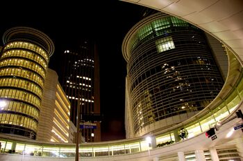 Night image of several tall skyscrapers taken from a street view, looking up. Several lights and traffic lights can be seen on the street, along with a round walkway above the street.