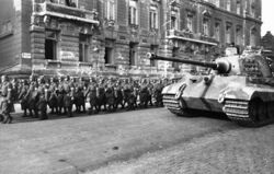 A large tank with sloped frontal armour and a flat faced turret, by a column of marching soldiers wearing overcoats and helmets, in a wide city street. A large building to the rear shows the scars of battle.