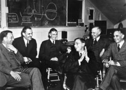 A group of in suits men sit around, laughing. On the blackboard behind is a discarded plan for assembling a uranium core.