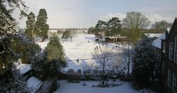 A panoramic vista of school playing fields covered in snow. Two large trees are visible to the left, schoolrooms to the right. A number of pupils are seen playing in the snow.