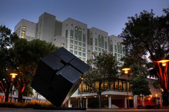 A dark gray sculpture of a tilted cube in front of a tower housing FIU's library