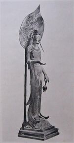 Three-quarter view of a very slim and tall statue carrying a vase in with two fingers of her left hand. Her right arm is bent with the palm of her right hand facing upward. A halo on a pole is seen behind the statue.