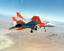 Jet aircraft with distinctive orange markings banking left over desert, with landing gears extended