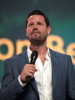 Portrait photograph of Seth Dillon from the shoulders up, wearing a white shirt and blue jacket, holding a microphone
