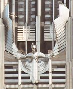 A stylized figure of a male human with outstretched arms and head tilted slightly forward, wearing a winged and crested helmet, mounted on the facade of a building
